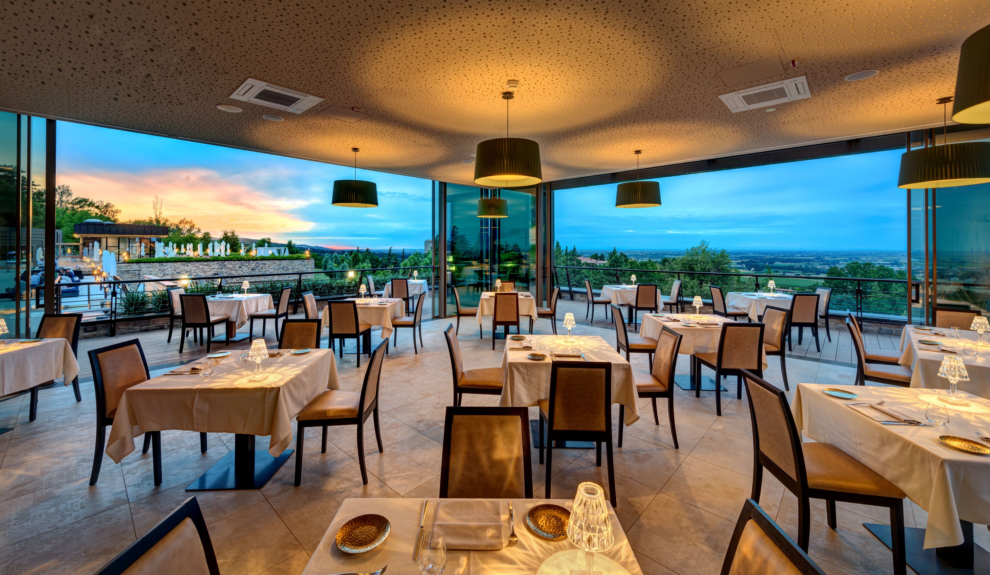 Un restaurante con mesas dispuestas para comer, amplias ventanas que ofrecen vistas al paisaje e iluminación colgante.