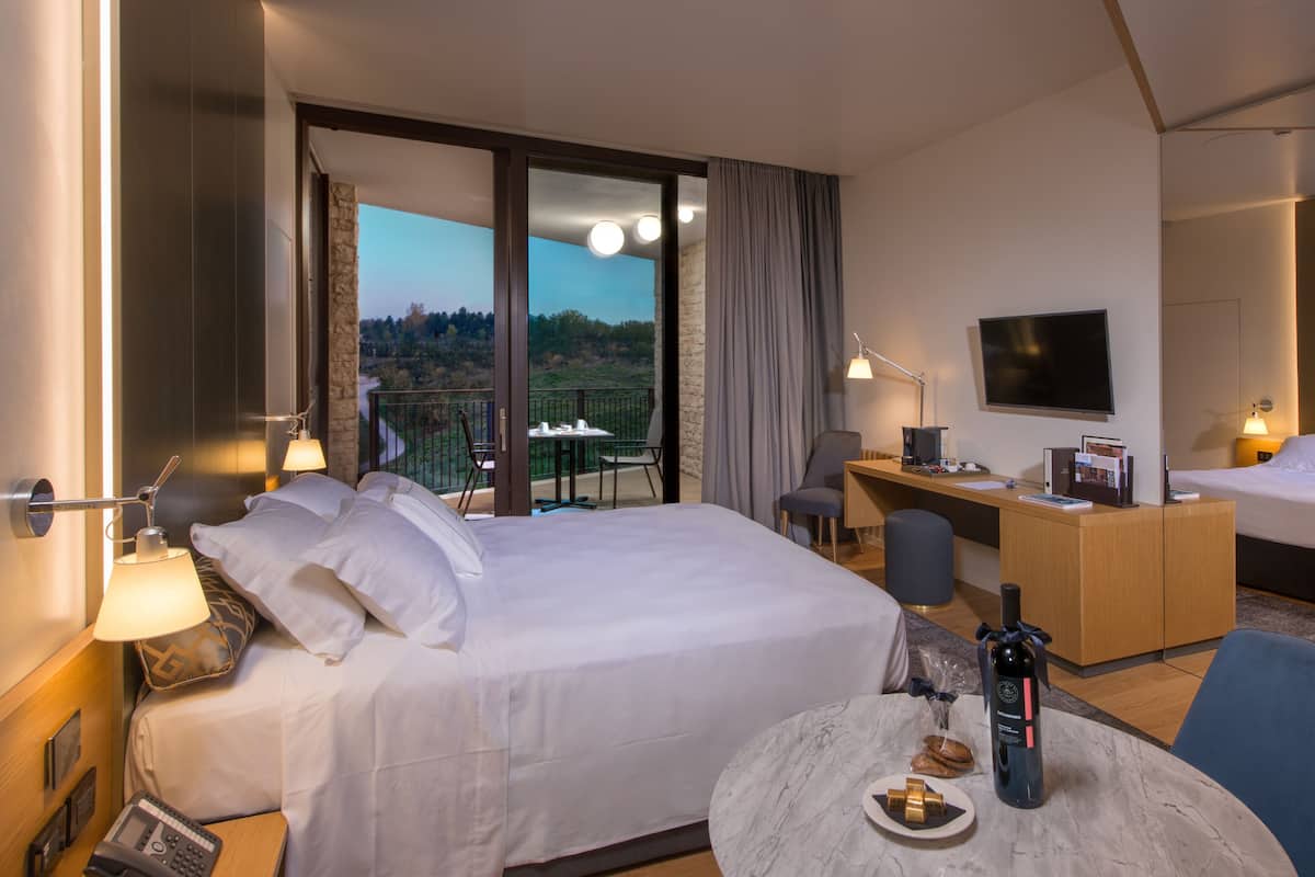 Ein Hotelzimmer mit einem großen Bett, einem Schreibtisch, einem Sessel, einem Fernseher und einem Balkon mit Blick auf die Landschaft.
