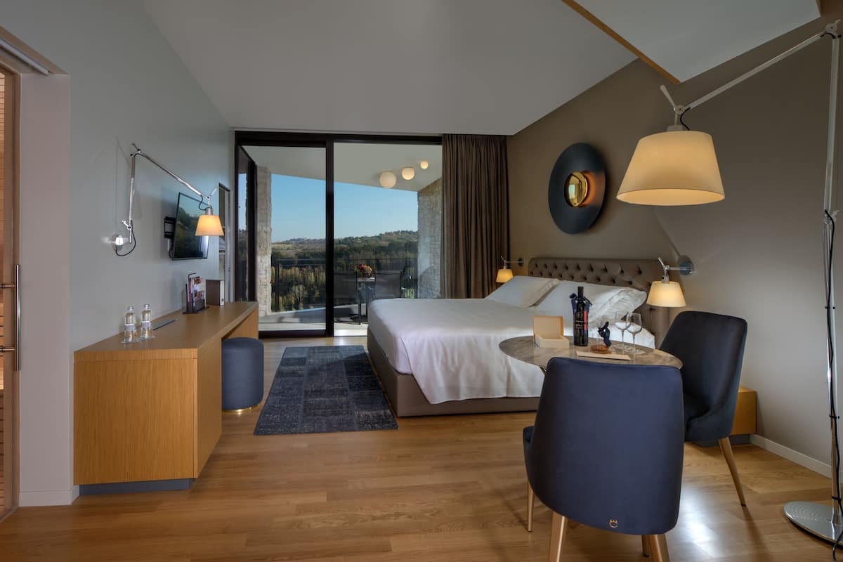 Ein modernes Hotelzimmer mit einem großen Bett, einem Schreibtisch, einem Sessel und Blick ins Freie.