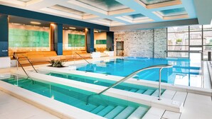 An indoor swimming pool with clear blue water, a wooden bench area, and a stone wall. - Palazzo di Varignana Resort & SPA (Castel San Pietro Terme)