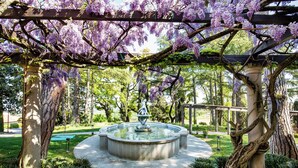 A garden with a fountain, trees, and a pergola covered with purple flowers. - Palazzo di Varignana Resort & SPA (Castel San Pietro Terme)