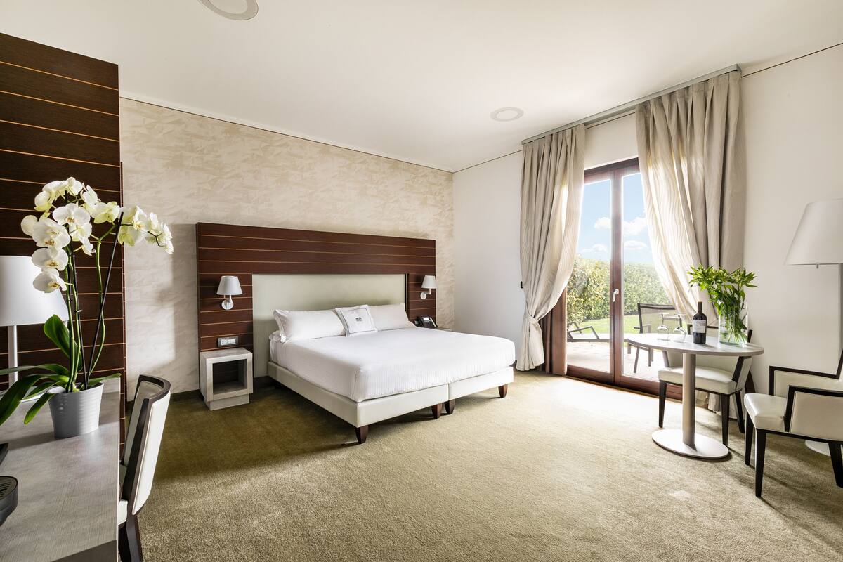 Ein Hotelzimmer mit Bett, Nachttischen, einem Schreibtisch mit Stuhl, einem Esstisch mit Stühlen und Blick ins Freie.