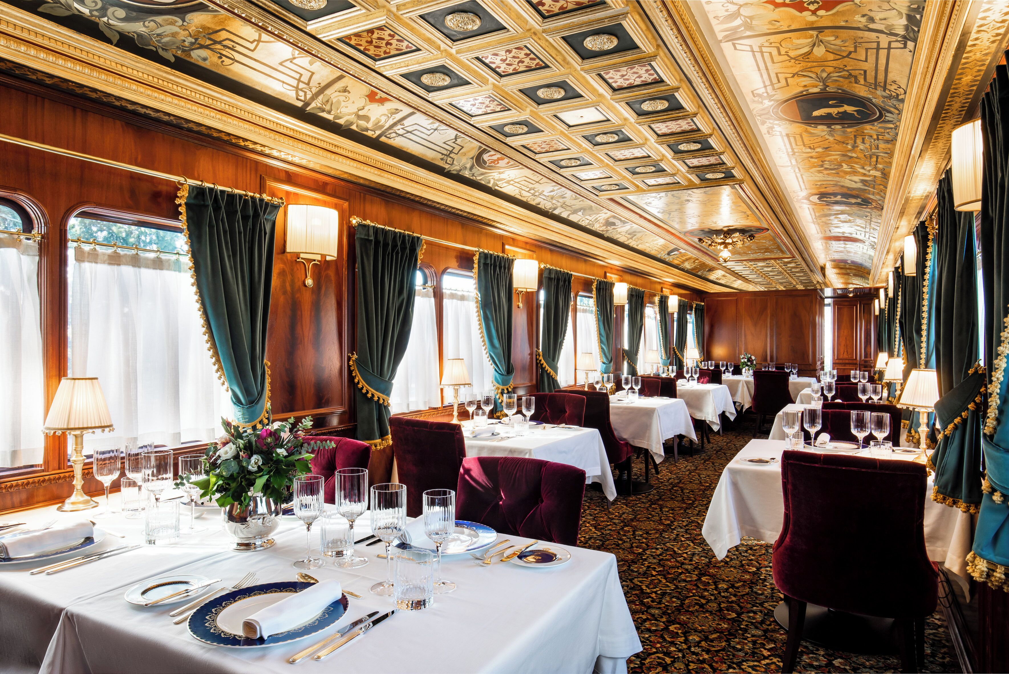 Un comedor lujoso con un techo ornamentado, elegantes mesas dispuestas y asientos mullidos.