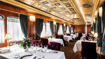 Un comedor lujoso con un techo ornamentado, elegantes mesas dispuestas y asientos mullidos.