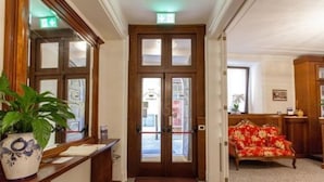 A hotel lobby with a reception desk, a plant, a red floral sofa, and a double door with a sign above it. - Hotel Garni Corona (Menaggio)