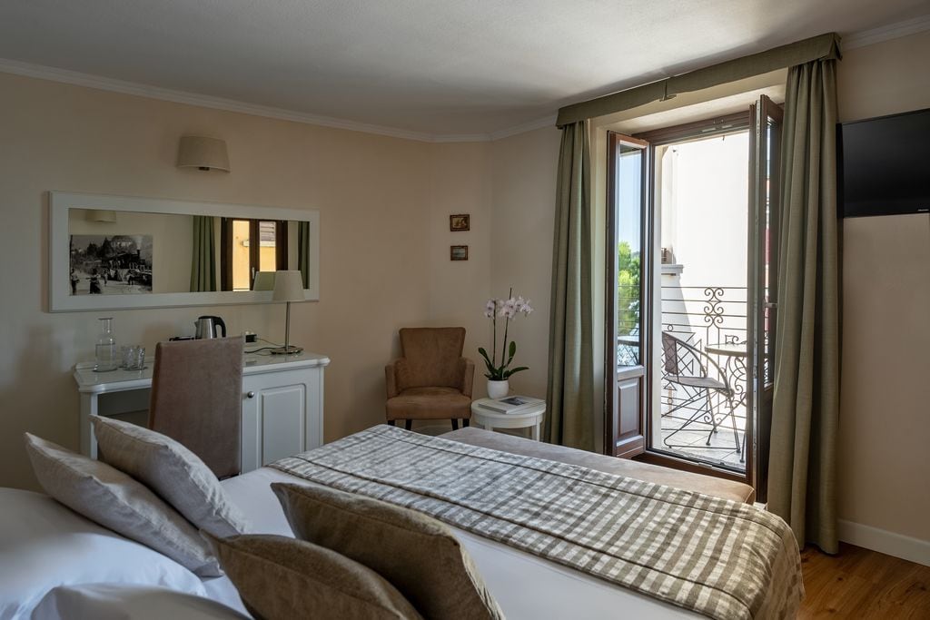 A hotel room with a bed, a desk, a chair, a small table with a lamp, and a balcony with a chair.