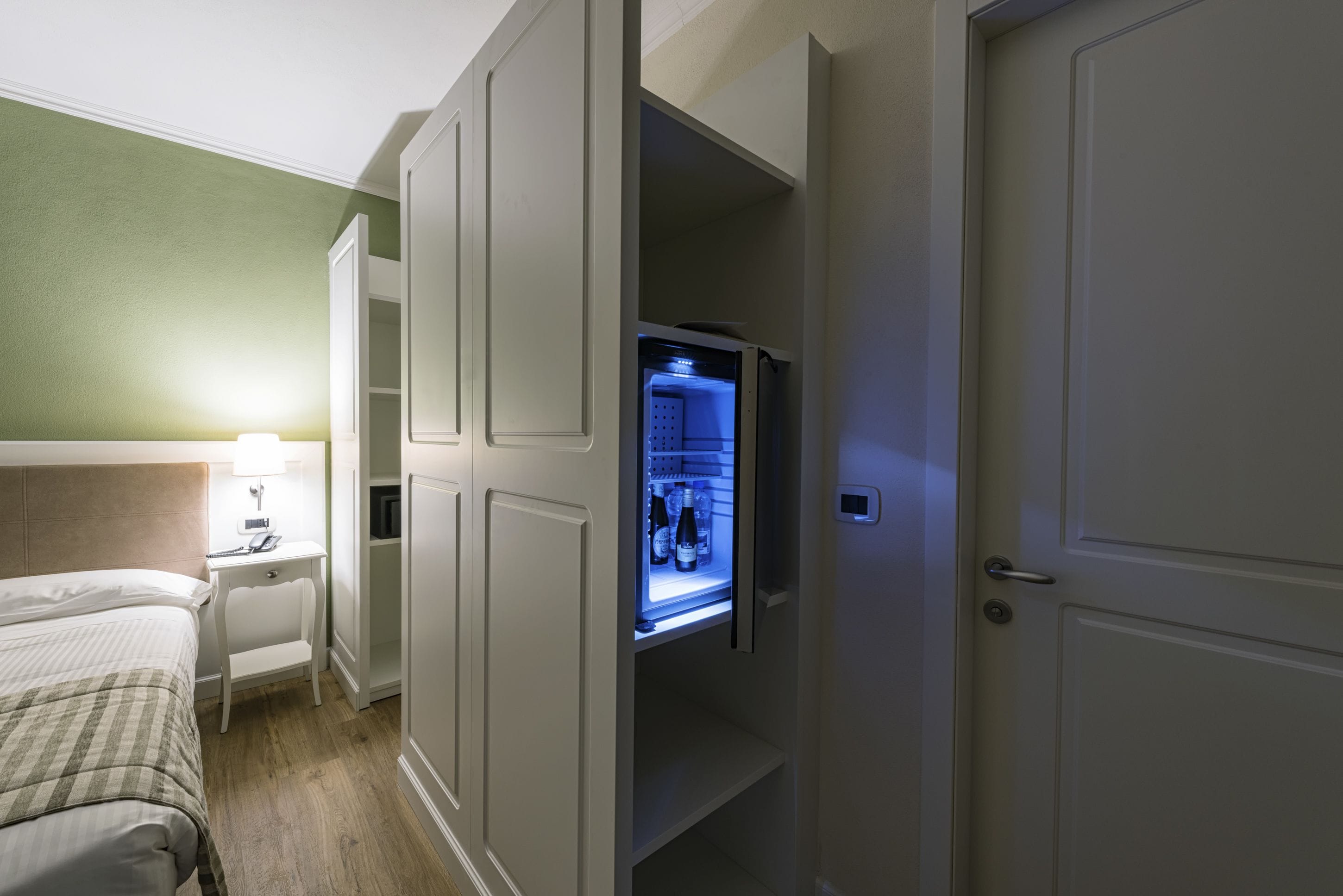 A hotel room with a bed, bedside table, lamp, and a built-in refrigerator.