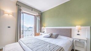 A hotel room with a large bed, a nightstand, a lamp, a window with curtains, and a view of the outdoors. - Hotel Garni Corona (Menaggio)