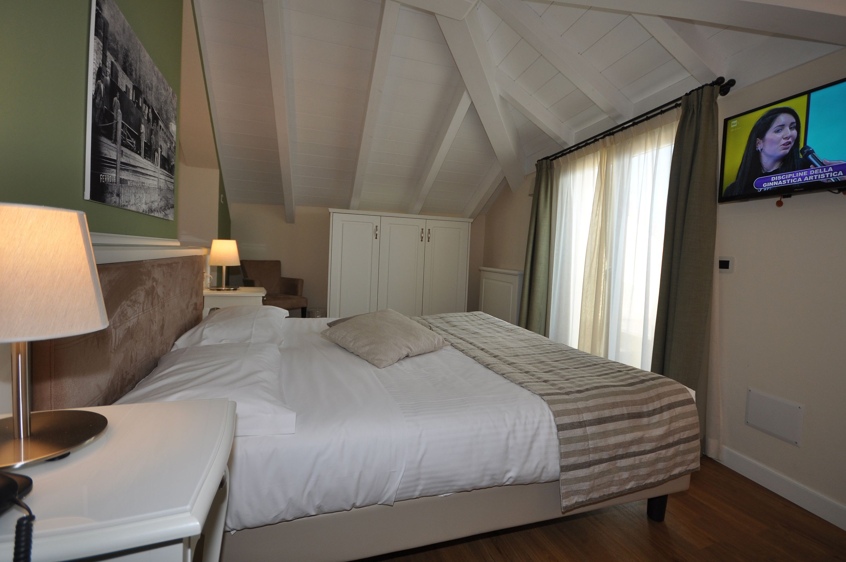 A bedroom with a bed, a nightstand, a lamp, a wardrobe, and a television on the wall.