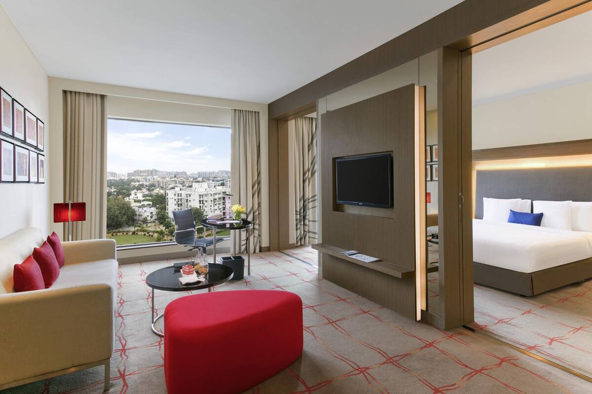 Une chambre d’hôtel avec un canapé, un pouf rouge, une télévision, un lit, une petite table avec un vase et une vue sur la ville.