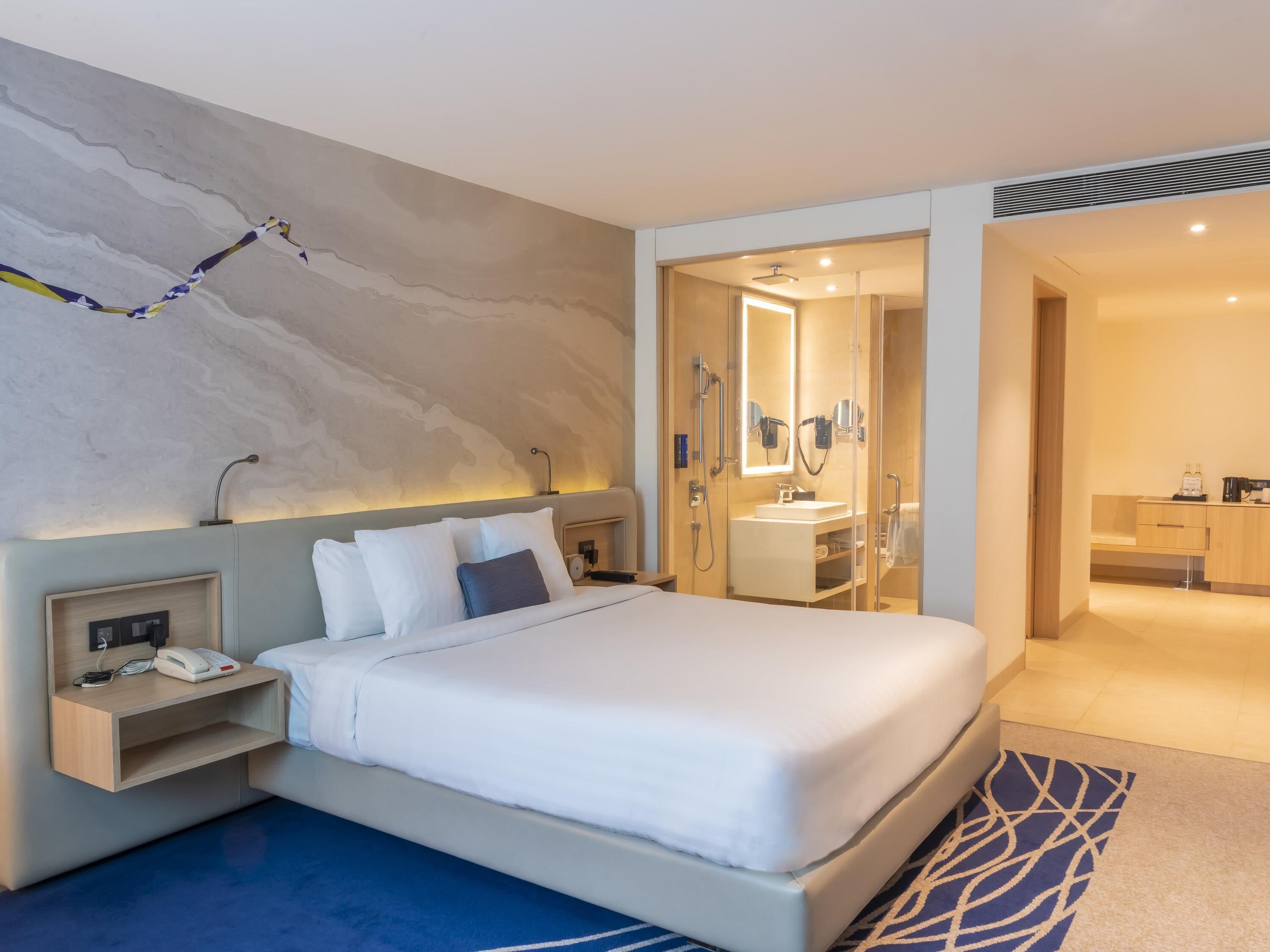 A modern hotel room with a large bed, bedside table with a phone, a mirror, and a shower area.