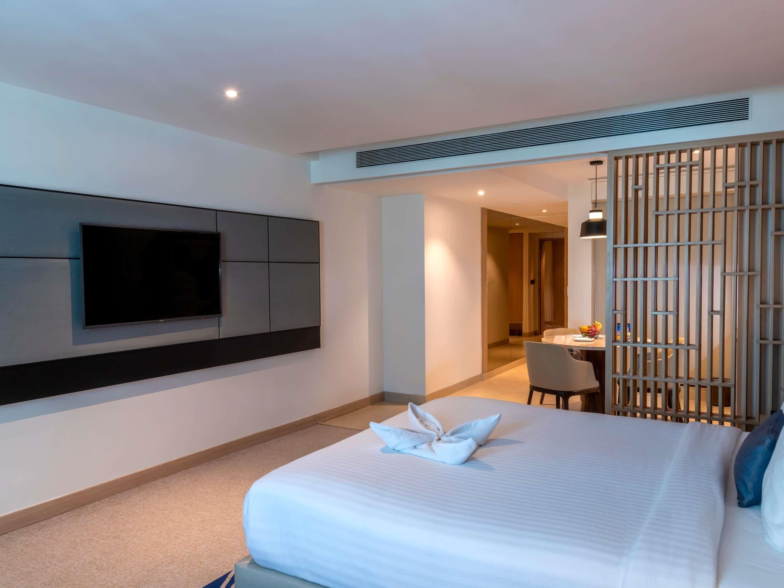 Une chambre d’hôtel moderne dotée d’un grand lit, d’un téléviseur à écran plat fixé au mur, d’un coin repas avec une table et des chaises, et d’une cloison décorative.