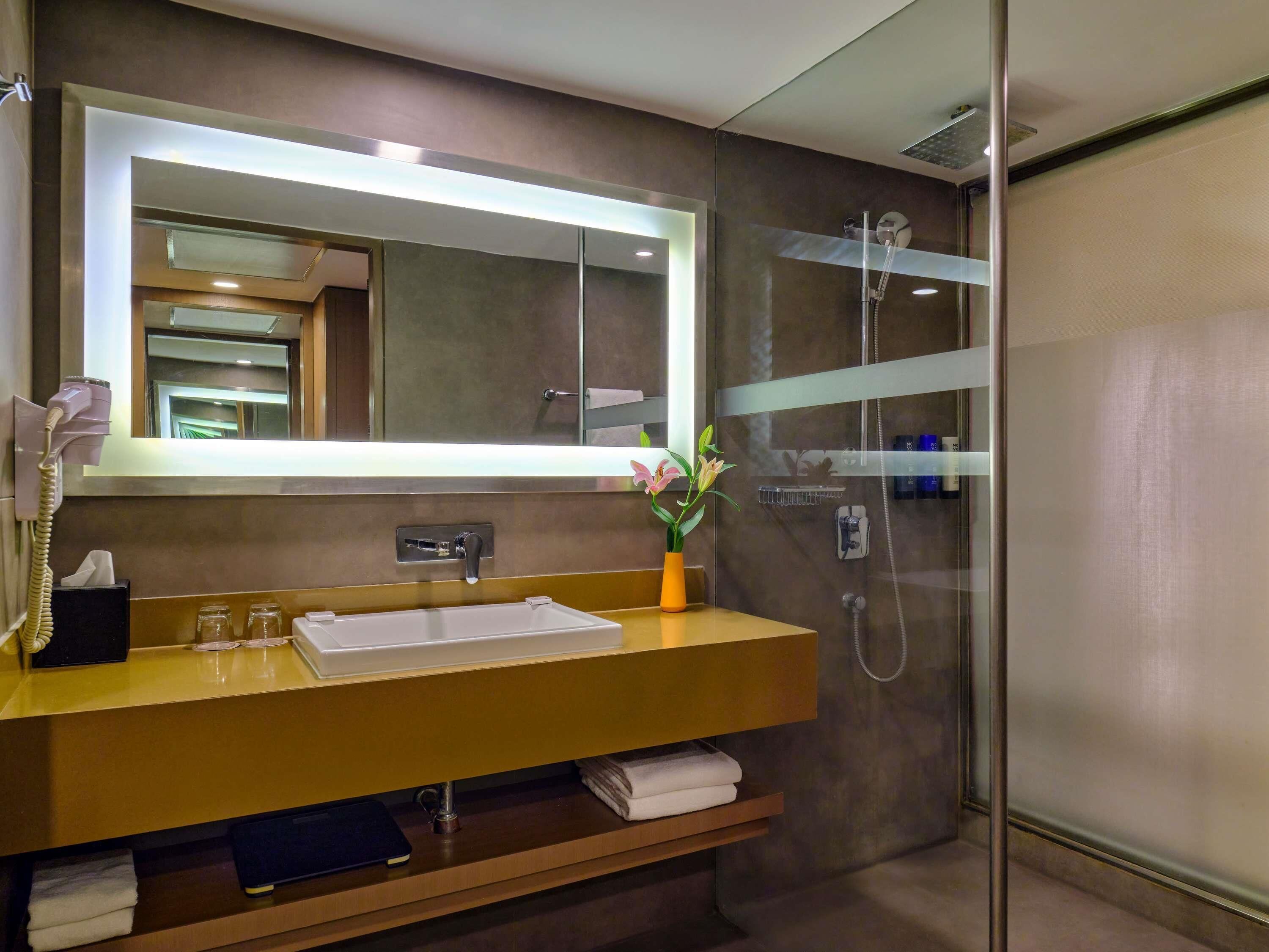 A modern bathroom with a large mirror, a sink with a towel rack, a glass shower enclosure, and a vase with flowers.