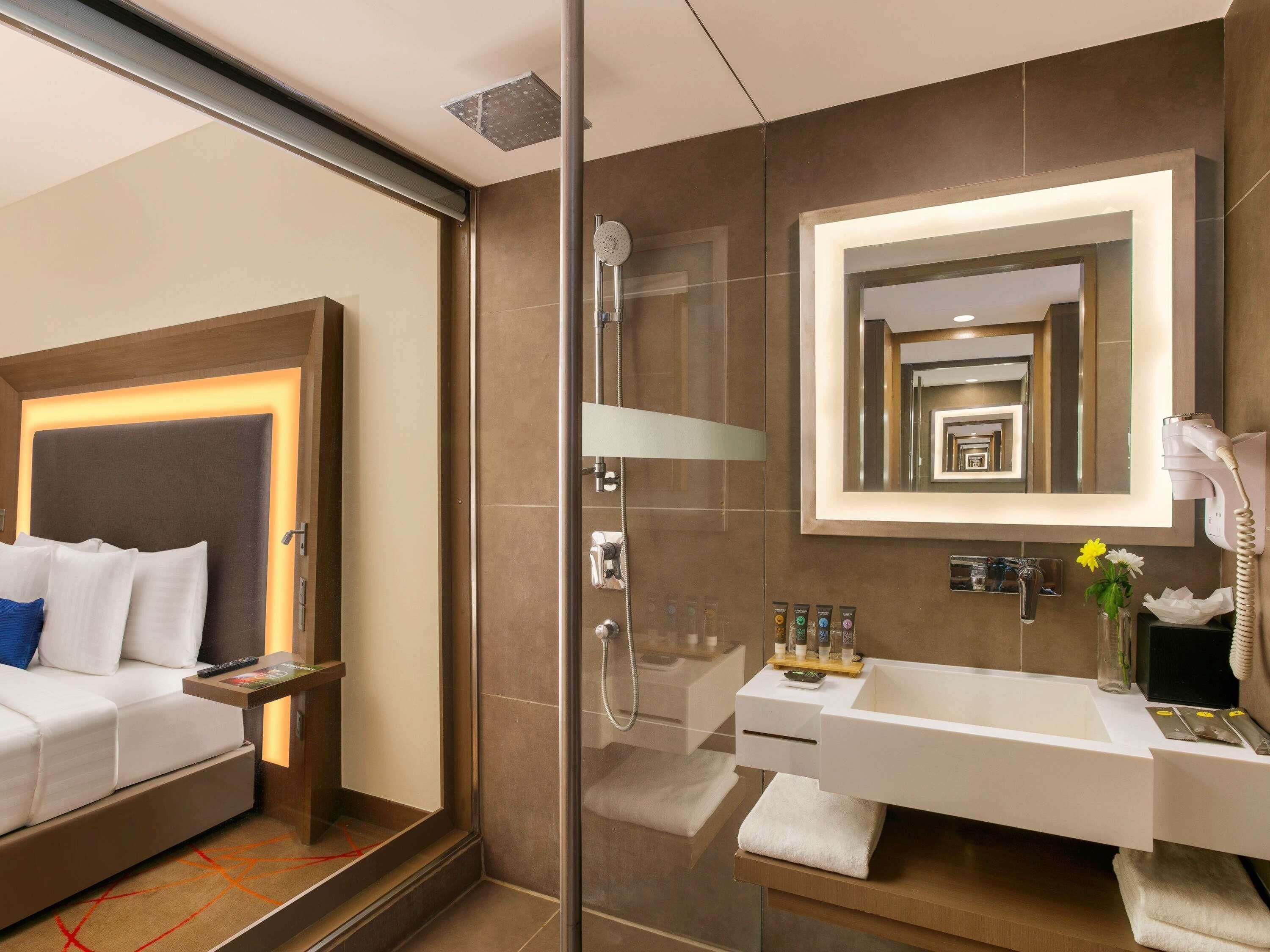 A modern hotel room with a bed, a glass shower enclosure, a large mirror, a vanity with a sink, and toiletries.