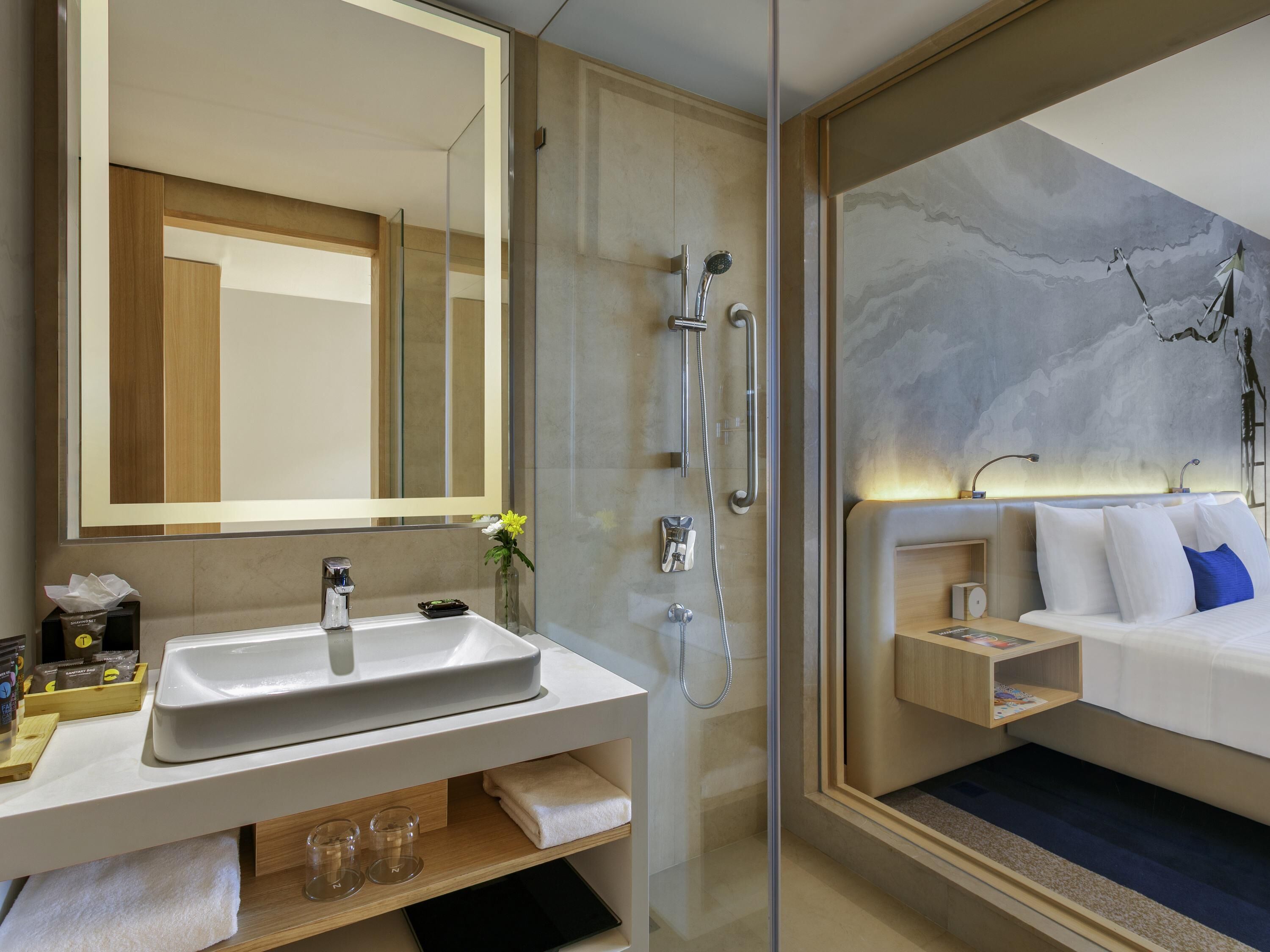 A modern bathroom with a glass shower enclosure, a white sink, and a bedside table with a lamp.