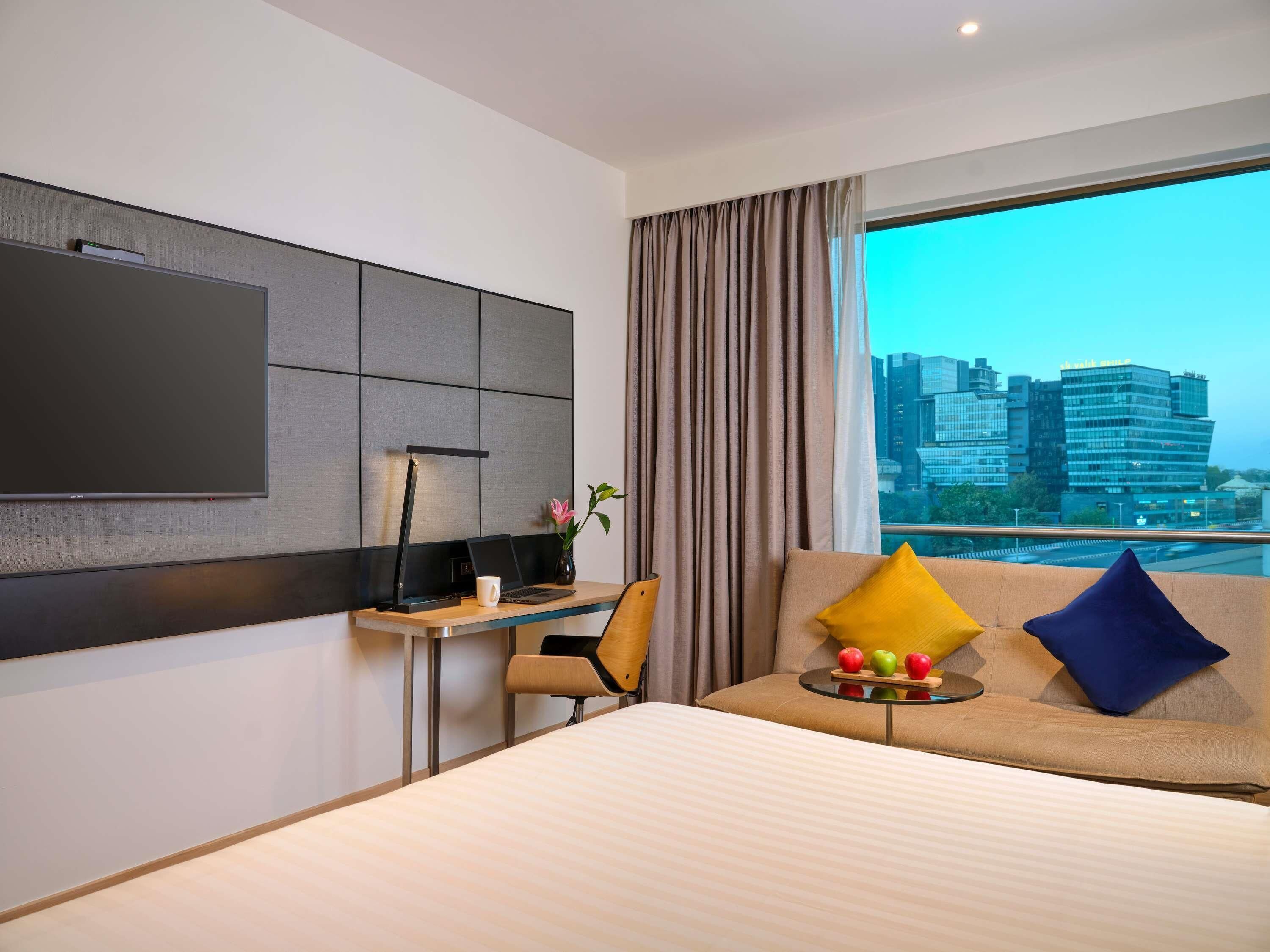 Une chambre d’hôtel moderne dotée d’un grand lit, d’un bureau avec un ordinateur portable, d’une télévision à écran plat et offrant une vue sur la ville par la fenêtre.