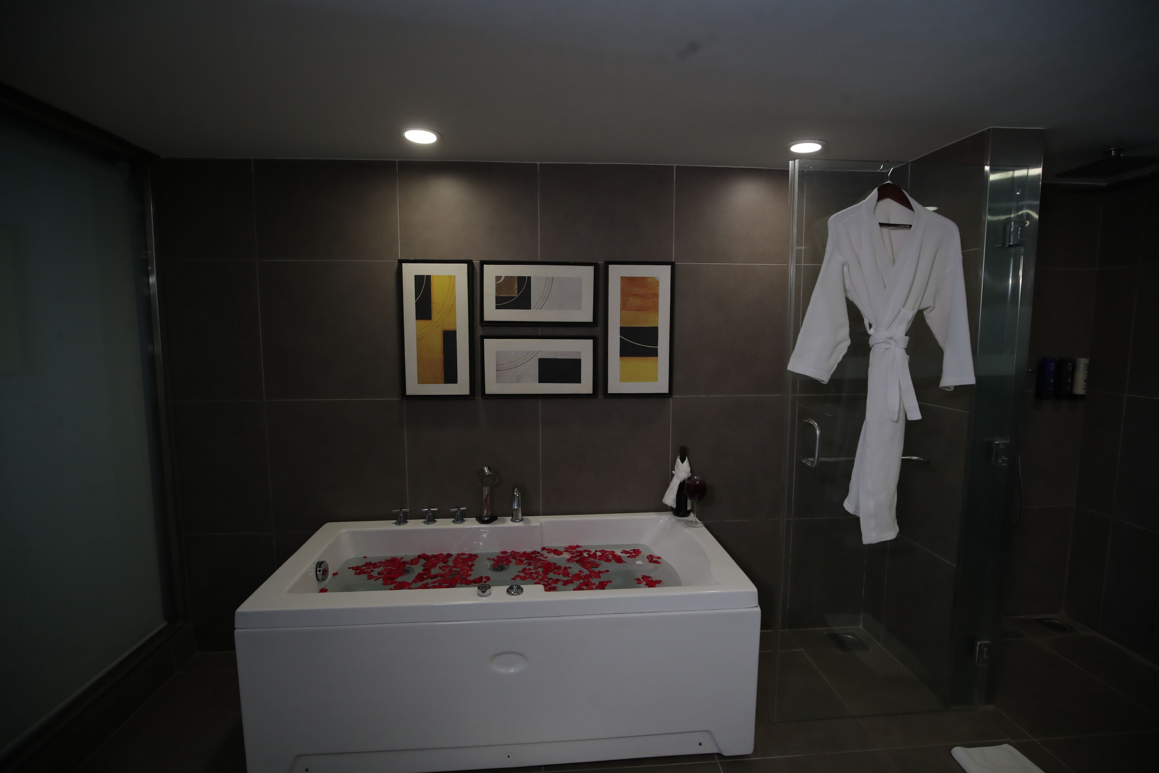 A modern bathroom with a large bathtub filled with rose petals, a glass shower enclosure, and a white robe hanging on the wall.