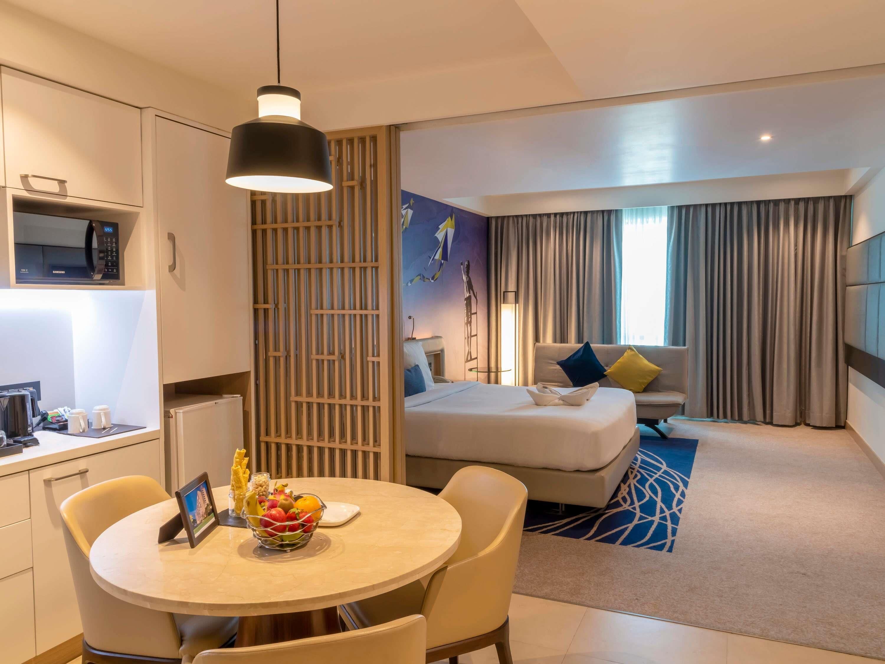 Une chambre d’hôtel moderne comprenant un lit, un coin repas avec une table et des chaises, un coin cuisine avec un four à micro-ondes et une grande fenêtre avec des rideaux.