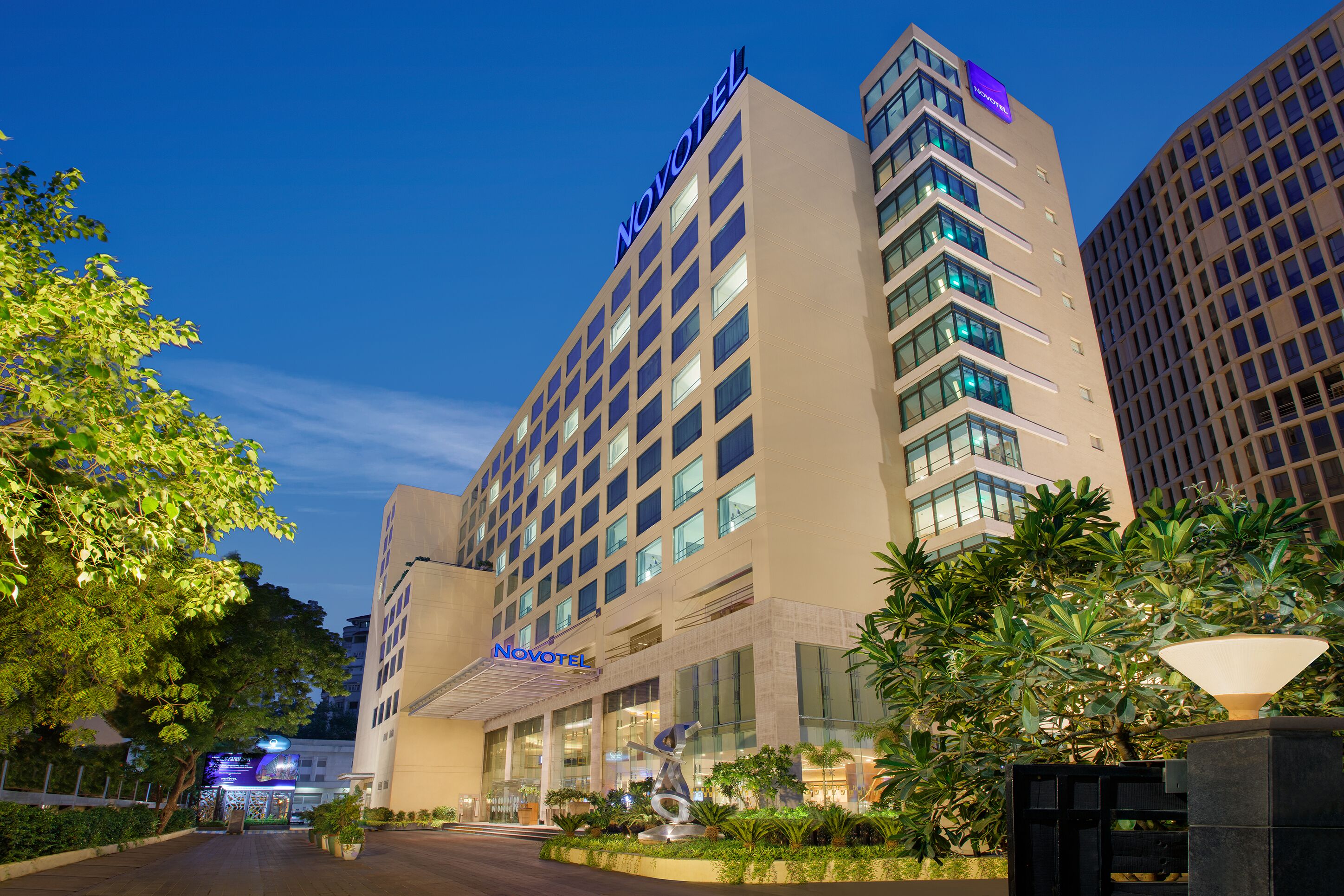 A hotel with a modern design, featuring a prominent 'Novotel' sign, a well-lit entrance, and a landscaped garden area.