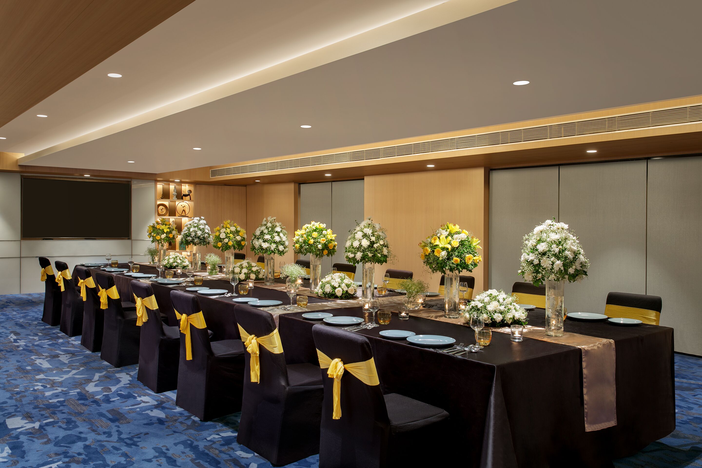 A conference room with long tables, chairs with yellow sashes, and floral centerpieces.