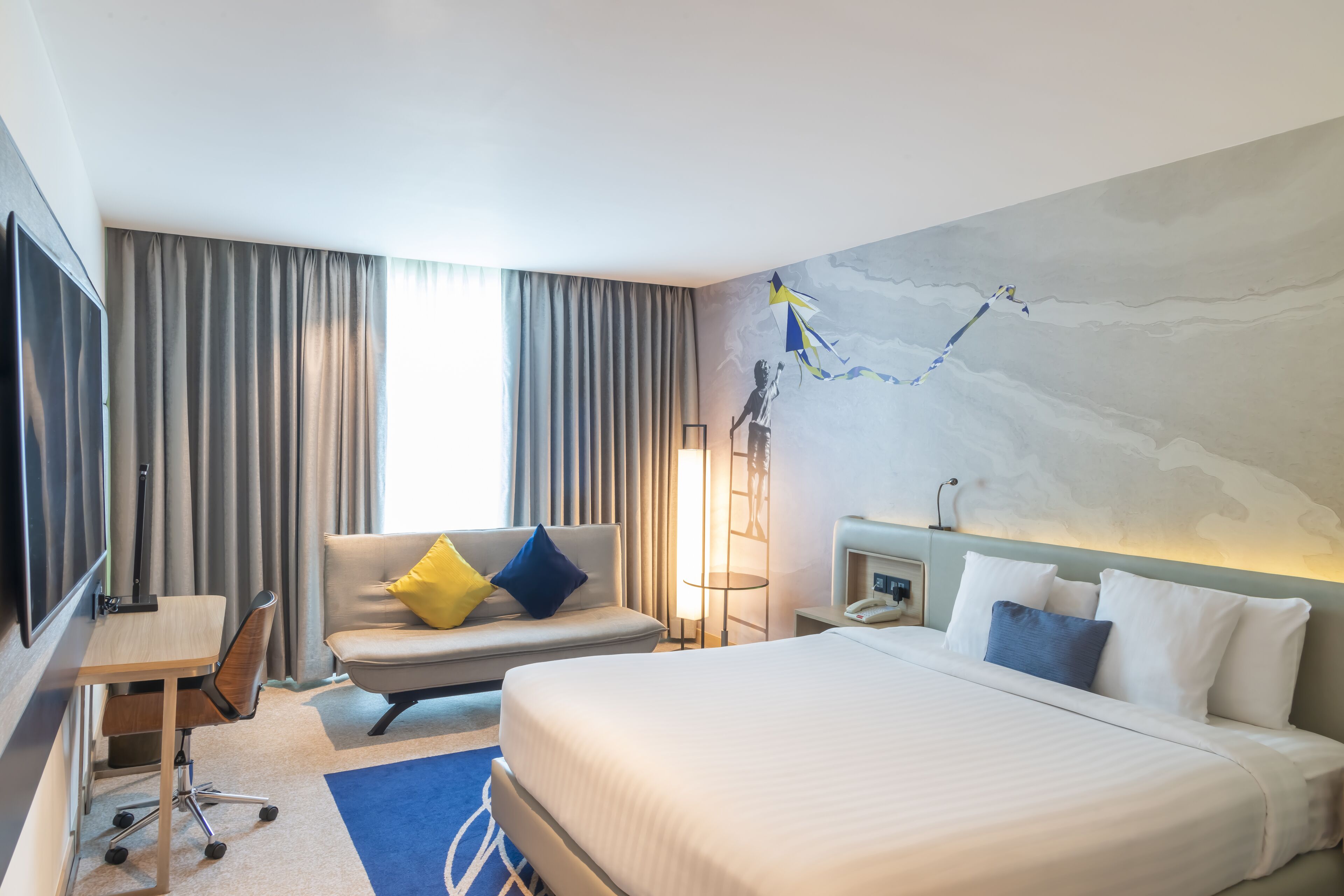 Une chambre d’hôtel comprenant un lit, un bureau, une chaise, un canapé avec des coussins, une lampe et un mur orné d’une fresque.