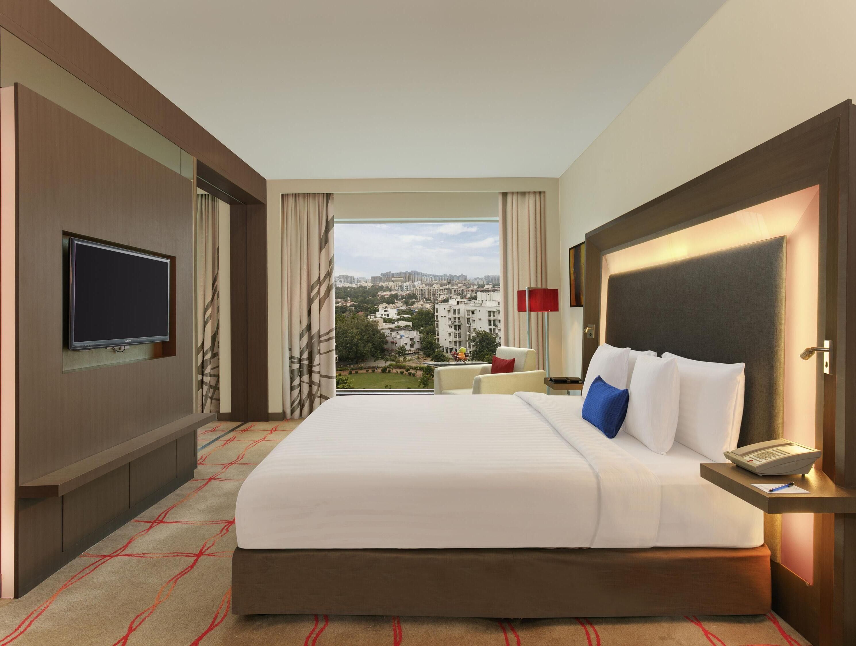 Habitación de hotel con una cama grande, televisión montada en la pared, vista a la ciudad y escritorio con lámpara.