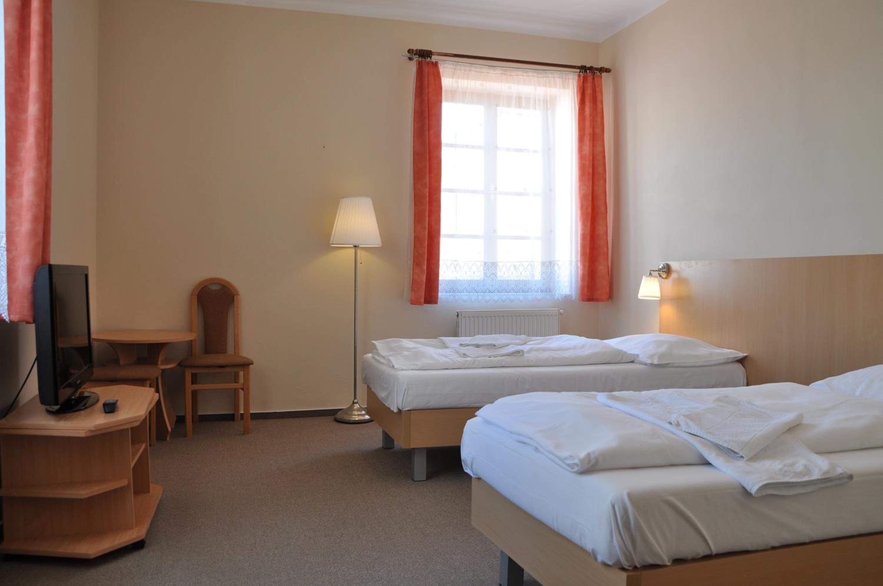 A hotel room with two beds, a desk, a chair, a TV, and a lamp.