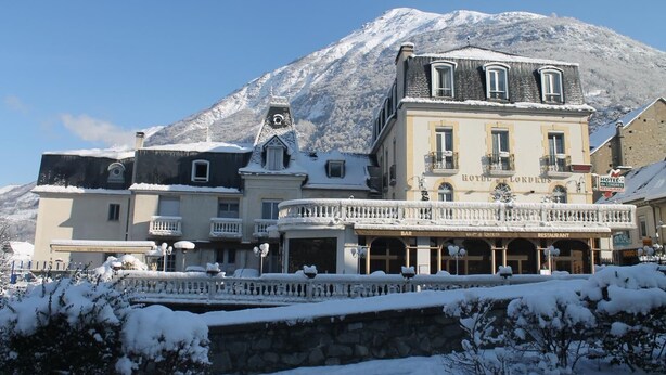 Hotel met een bar en restaurant op de begane grond, een balkon met sneeuw en een berg op de achtergrond.