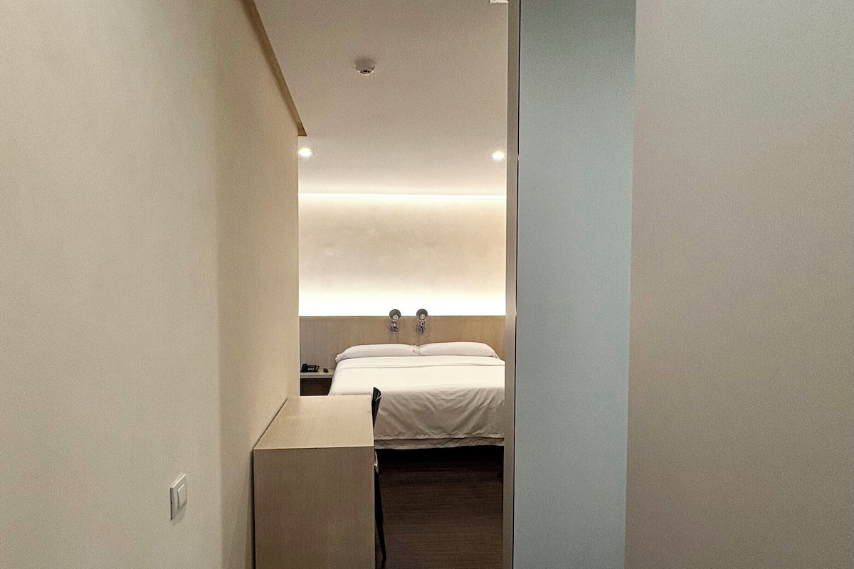 Ein schmaler Flur führt zu einem modernen Schlafzimmer mit Bett, Nachttisch und Wandleuchte.
