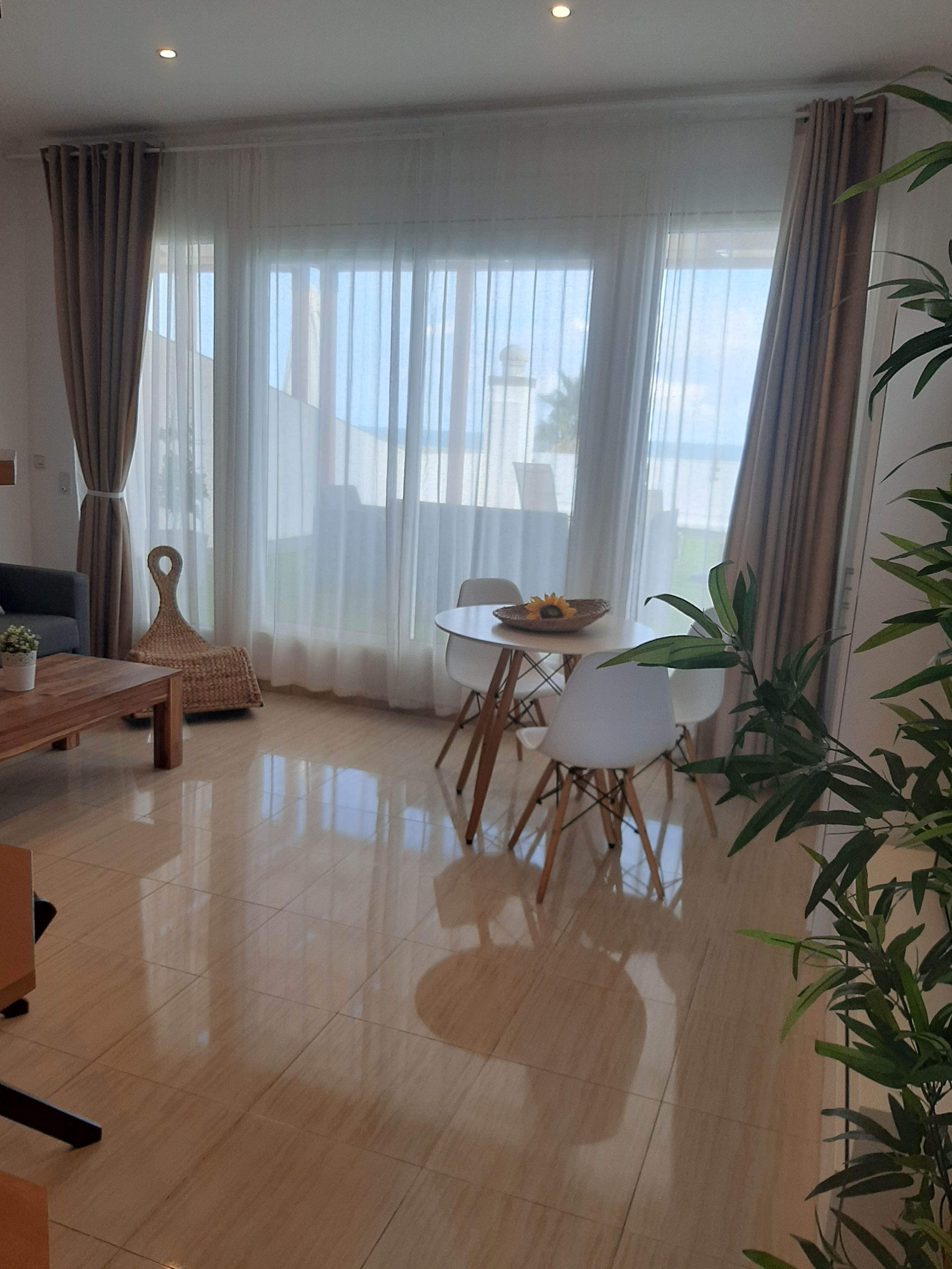 Ein modernes Wohnzimmer mit Essbereich, großen Fenstern mit durchscheinenden Vorhängen und Blick ins Freie.