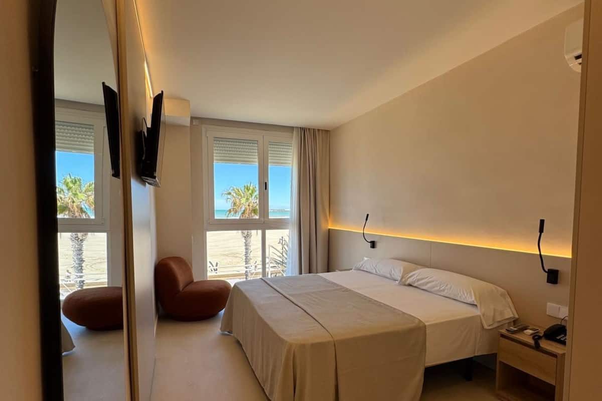 Ein modernes Hotelzimmer mit einem großen Bett, einem Nachttisch mit Lampe, einem Sessel und einem großen Spiegel.