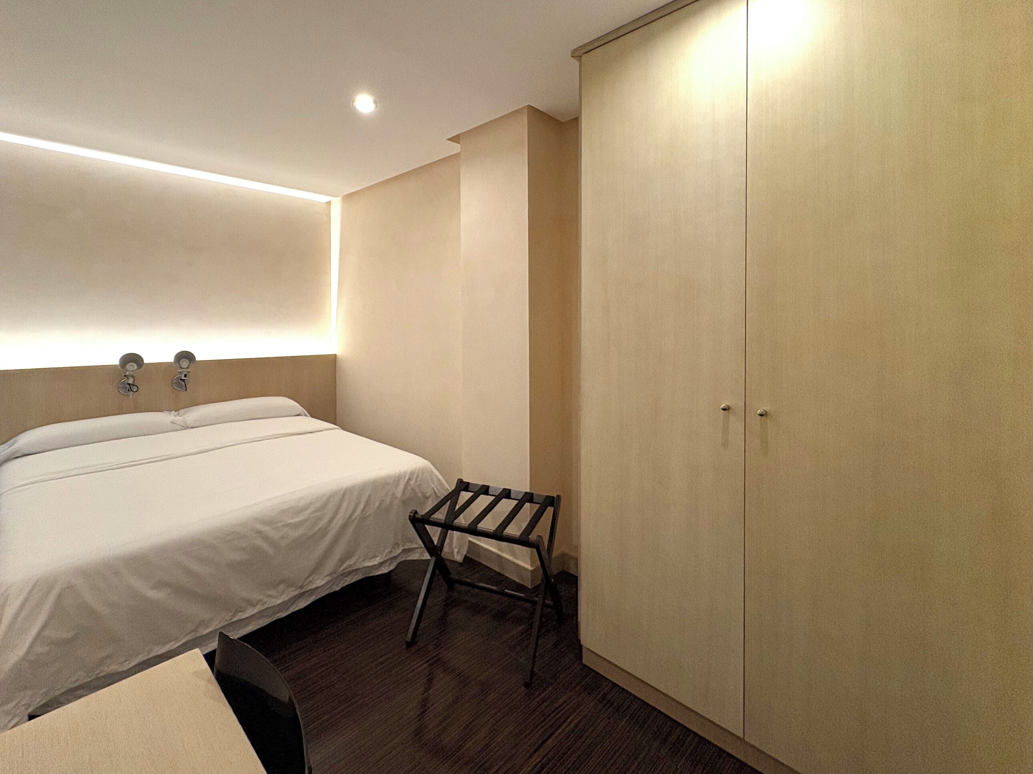 A hotel room with a bed, a chair, a wardrobe, and a wall-mounted light fixture.