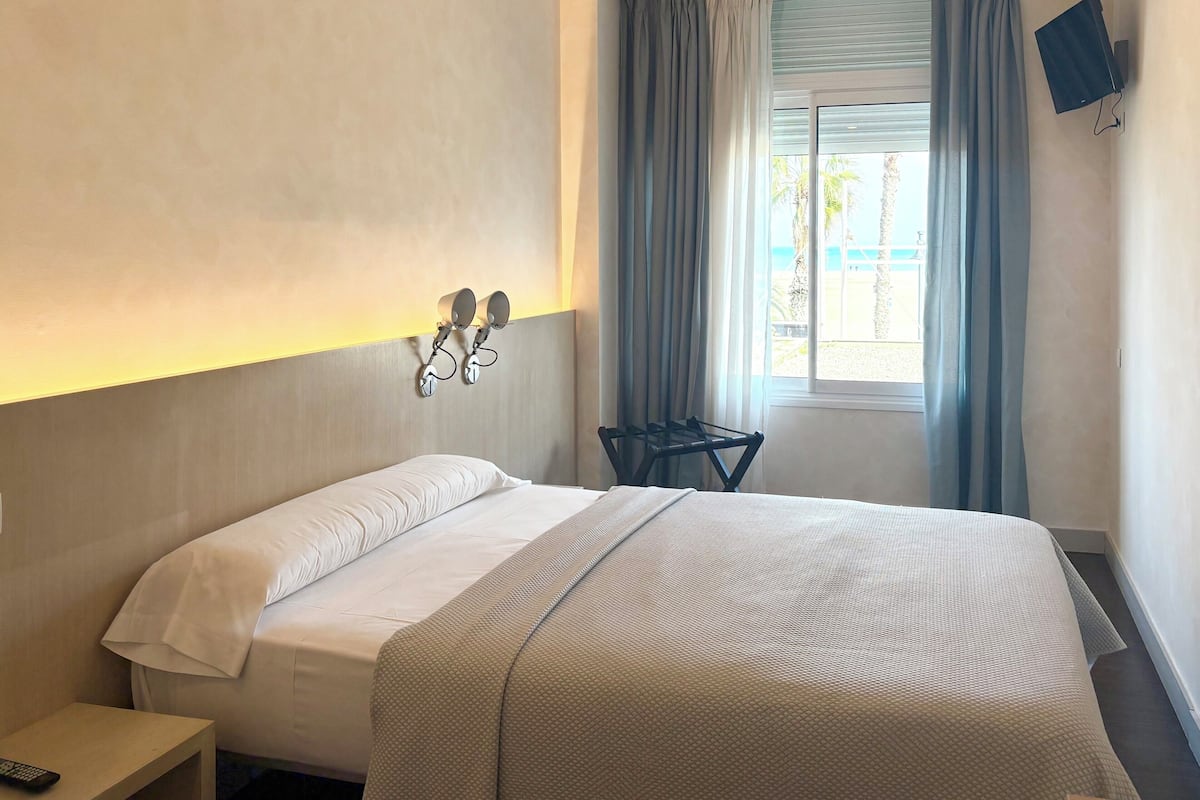 Ein Hotelzimmer mit einem Bett, einem Nachttisch mit Fernbedienung, einem an der Wand montierten Fernseher und einem Fenster mit Vorhängen.