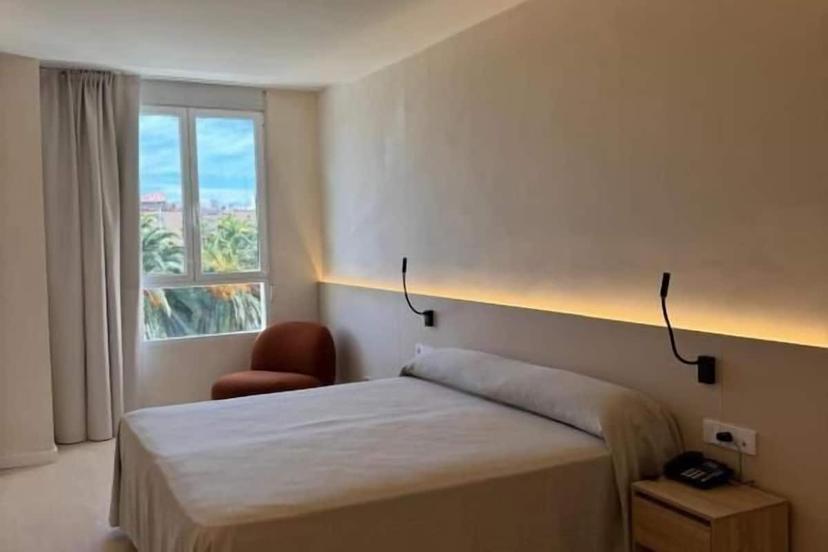 Ein modernes Hotelzimmer mit einem großen Bett, einem Sessel, einem Nachttisch und einem Fenster mit Blick auf Palmen.