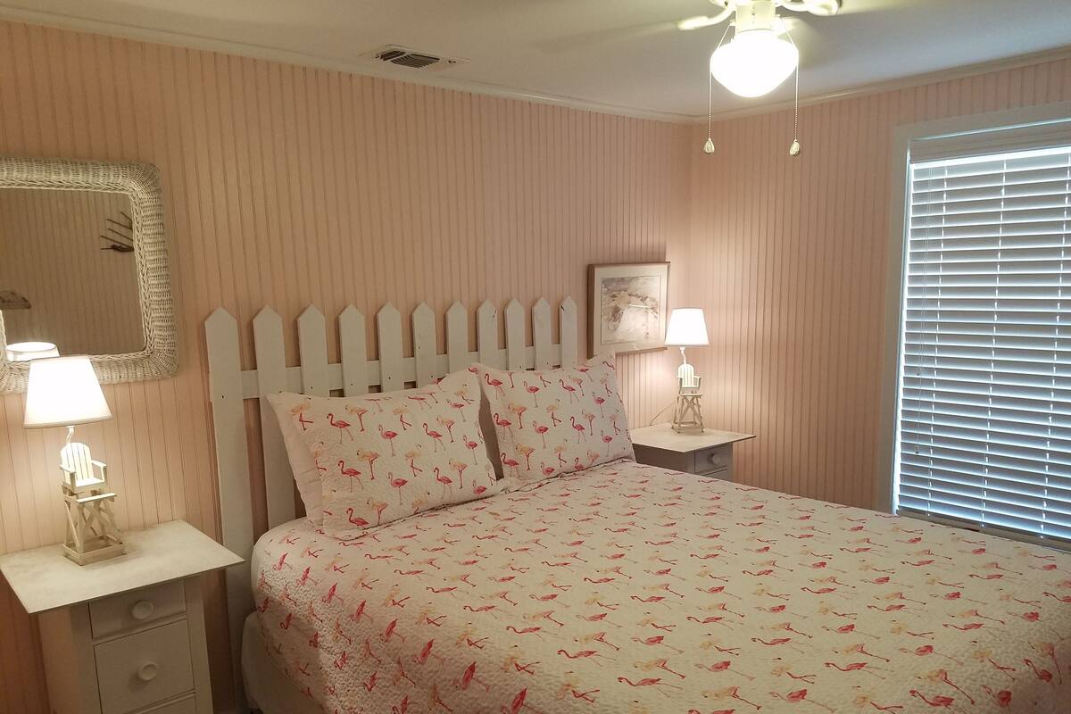 Une chambre à coucher comprenant un lit, deux tables de chevet avec des lampes, un miroir fixé au mur, un ventilateur de plafond et une fenêtre avec des stores.