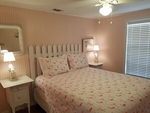 A bedroom with a bed, two bedside tables with lamps, a wall-mounted mirror, a ceiling fan, and a window with blinds.