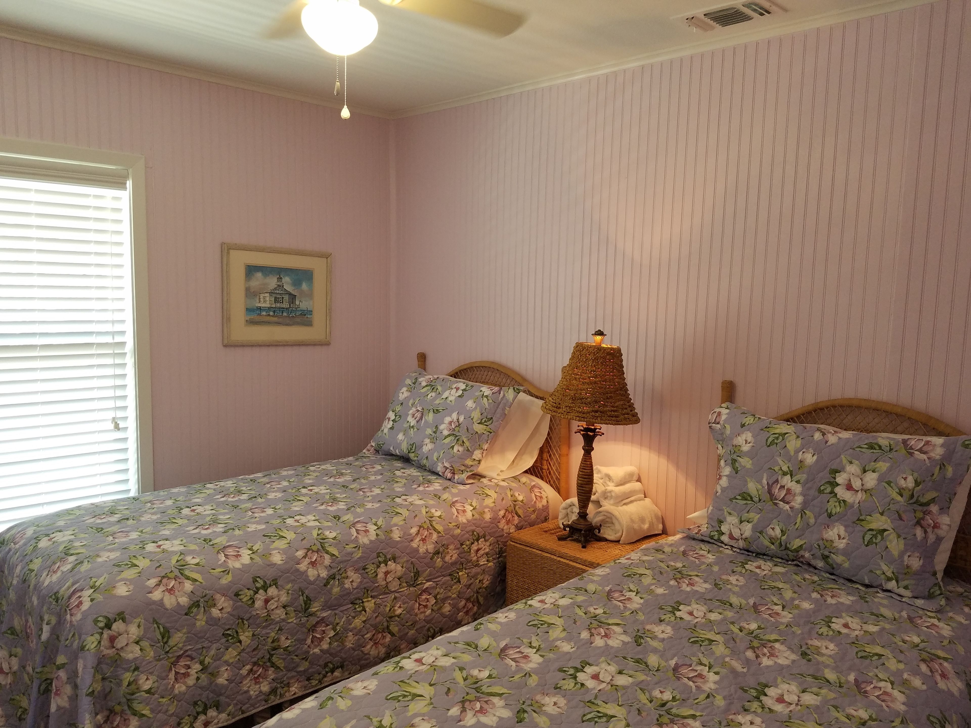 Une chambre avec deux lits, une table de chevet, une lampe, un ventilateur de plafond et une fenêtre avec des stores.