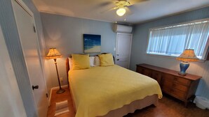 A bedroom with a bed, a ceiling fan, a night sky painting, a window with blinds, and a nightstand with a lamp.