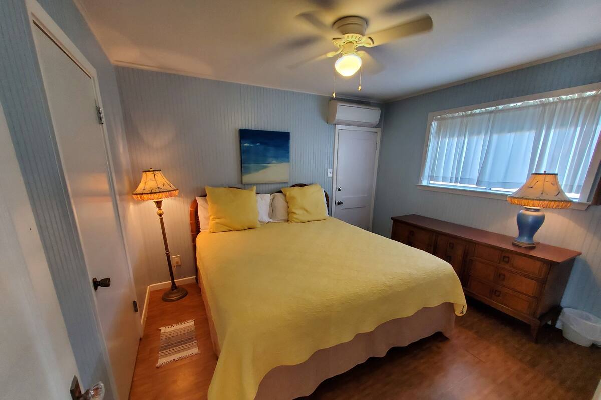 Une chambre à coucher avec un lit, un ventilateur de plafond, un tableau représentant un ciel étoilé, une fenêtre avec des stores et une table de chevet avec une lampe.