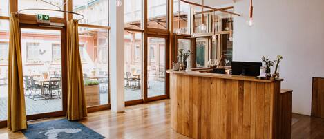 Un mostrador de recepción de madera en una habitación luminosa con ventanas amplias, un tapete azul y lámparas colgantes.