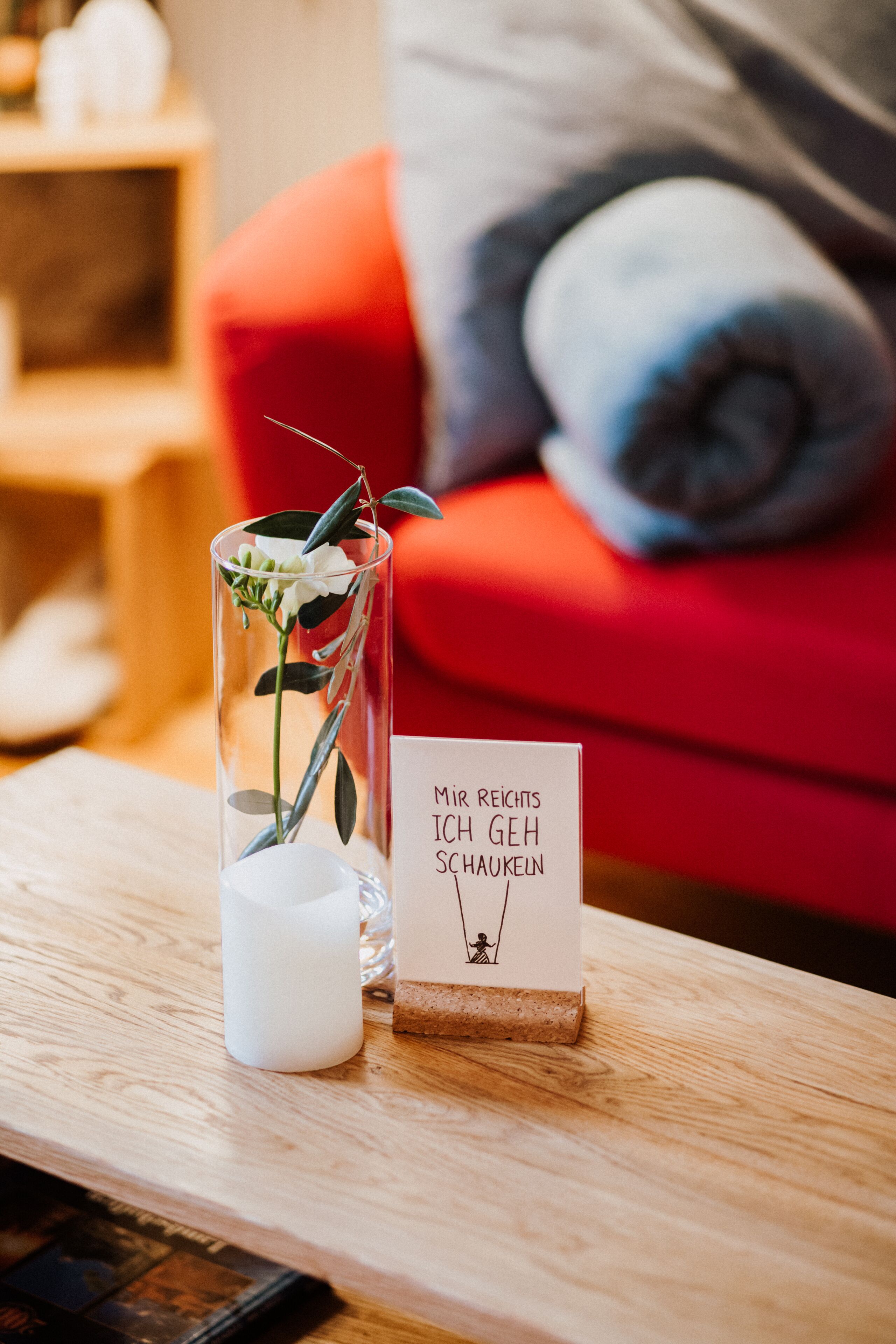 A clear glass vase with a white flower on a wooden table, a small card with text, and a red couch in the background.