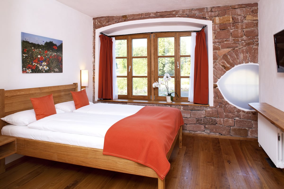 Een slaapkamer met een houten bed, een stenen muur, een raam met gordijnen en een schilderij aan de wand.