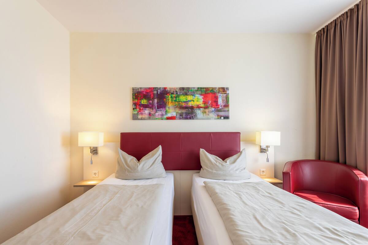 Habitació d'hotel amb dos llits, un capçal vermell, un quadre abstracte i colorit, i una butaca vermella.