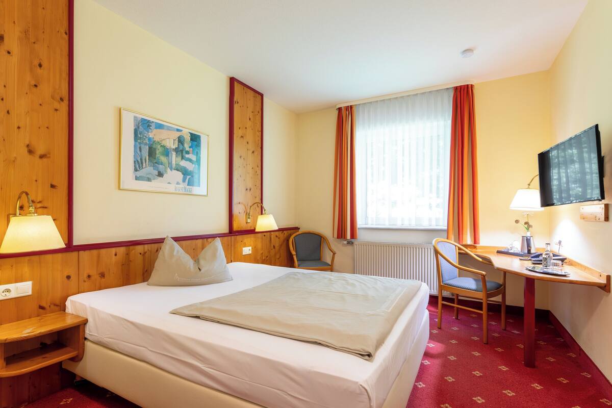 Una habitació d'hotel amb un llit, dues làmpades de nit, un escriptori amb una cadira, una televisió i una finestra amb cortines.