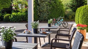 Un café al aire libre con sillas y mesas de mimbre, plantas en macetas y una bicicleta al fondo.