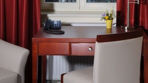 Habitación de hotel con un escritorio de madera, una silla, una lámpara y una ventana con cortinas rojas.