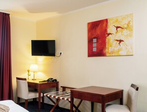 A hotel room with a bed, a desk with a chair, a television mounted on the wall, and a painting. - AppartementHotel SchlafGut Leipzig (Leipzig)