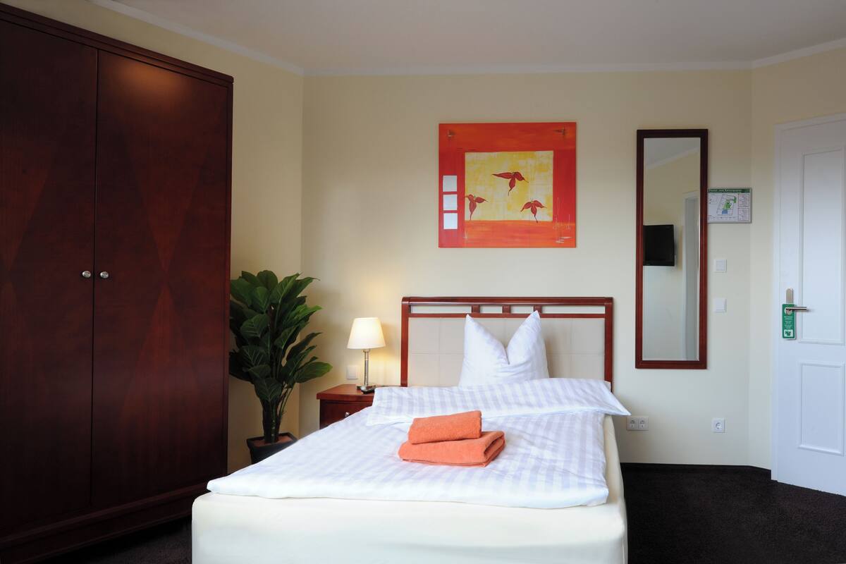 A hotel room with a large wardrobe, a bed with white bedding, a nightstand with a lamp, a potted plant, and a wall art piece.