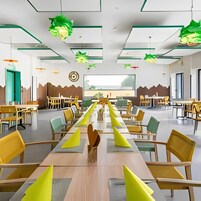 Un comedor amplio con mesas y sillas largas, luces colgantes verdes y vistas al exterior.