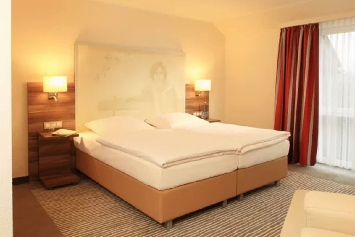A hotel room with a large bed, two bedside tables, a nightstand, a window with curtains, and a wall-mounted lamp.